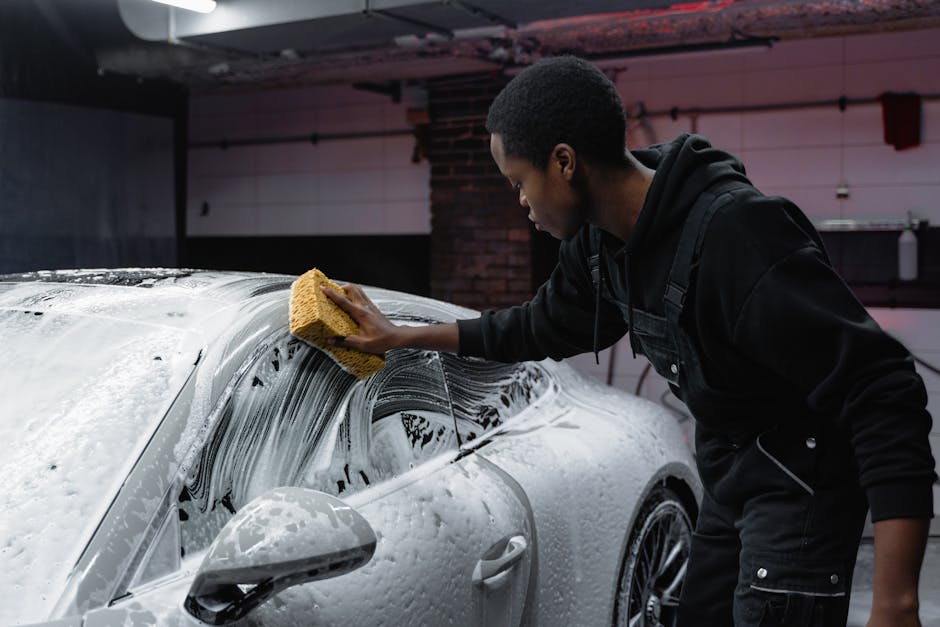 A young man cleans a luxury car with a sponge in an indoor carwash setting.